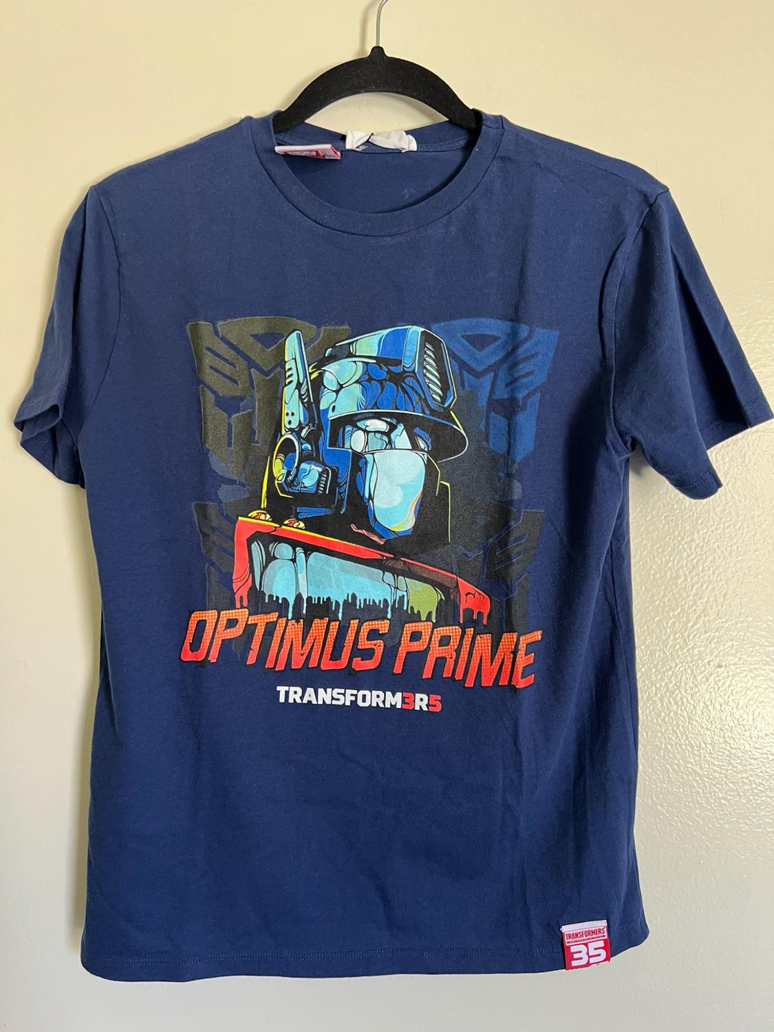 Blue T-shirt hanging on a hanger against a light background. Robot graphic and text OPTIMUS PRIME TRANSFORM3R5. Label TRANSFORMERS 35.