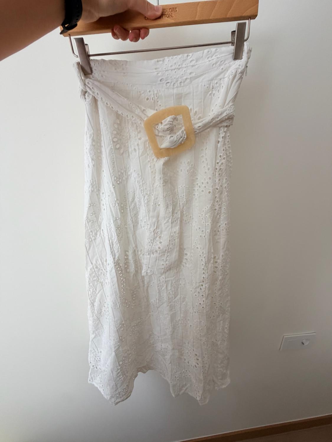 Hand holding up a white Swiss embroidery skirt with a square buckle belt in front of a white wall.