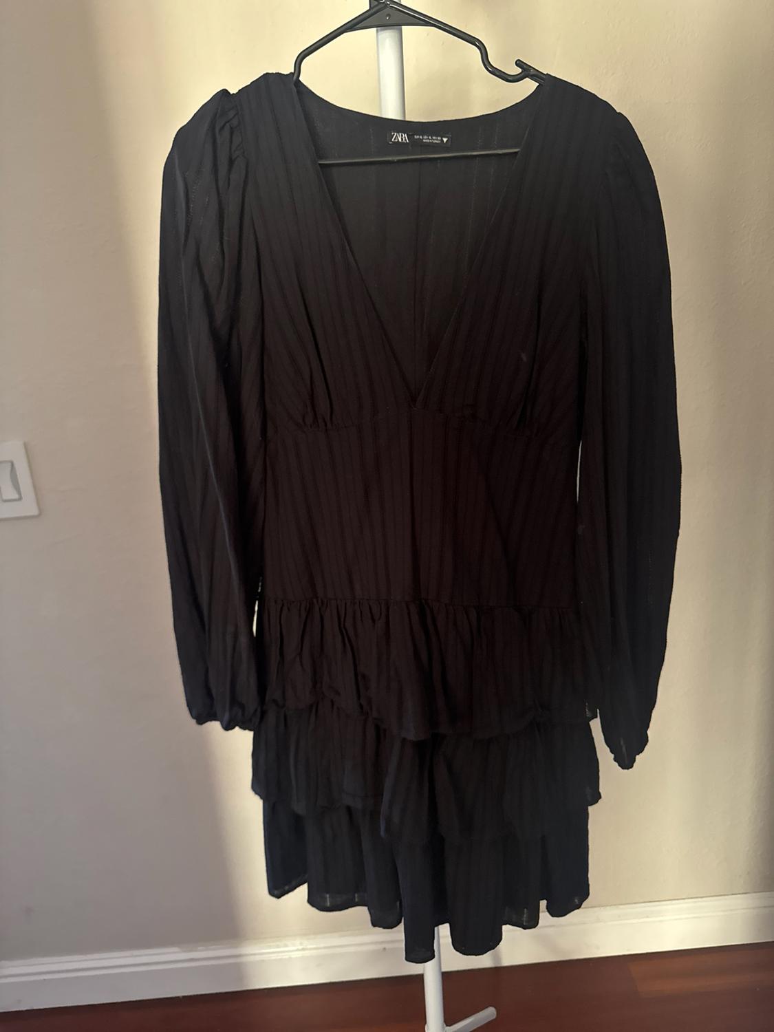 Long-sleeve black dress with a V-neck and ruffled skirt, hanging on a hanger against a light-colored wall. ZARA label.