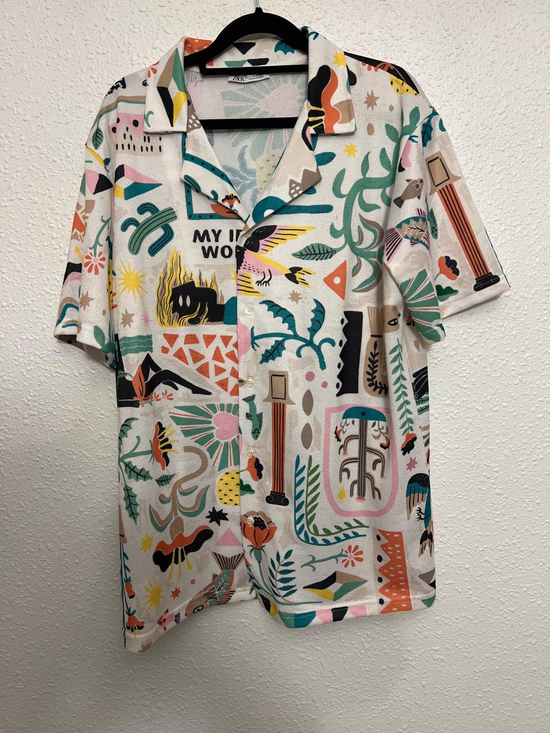 Colourful shirt with abstract prints and text "MY IN WO" hanging on a black hanger against a white wall.