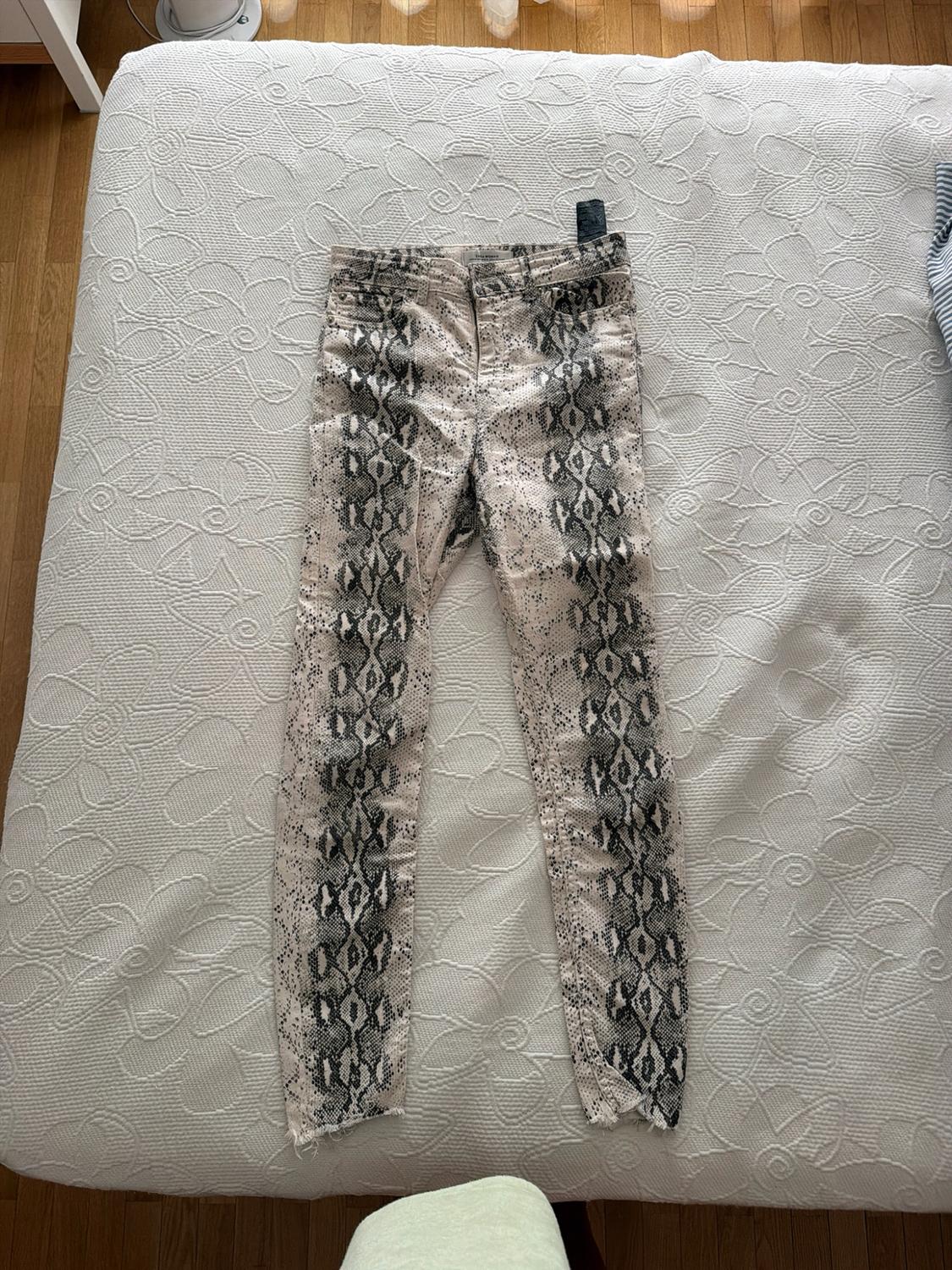 Snake-print trousers with frayed hems, laid out on a bed with a white textured bedspread. Casual and relaxed style.