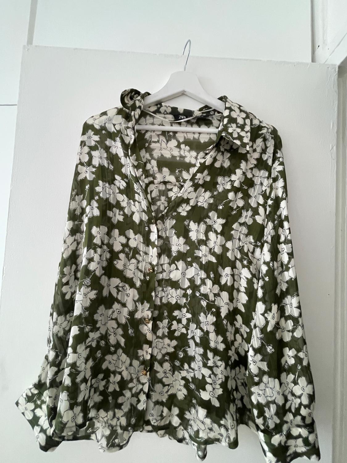 Green long-sleeved shirt with a white floral print, hanging on a hanger against a white door. The label reads ZARA.