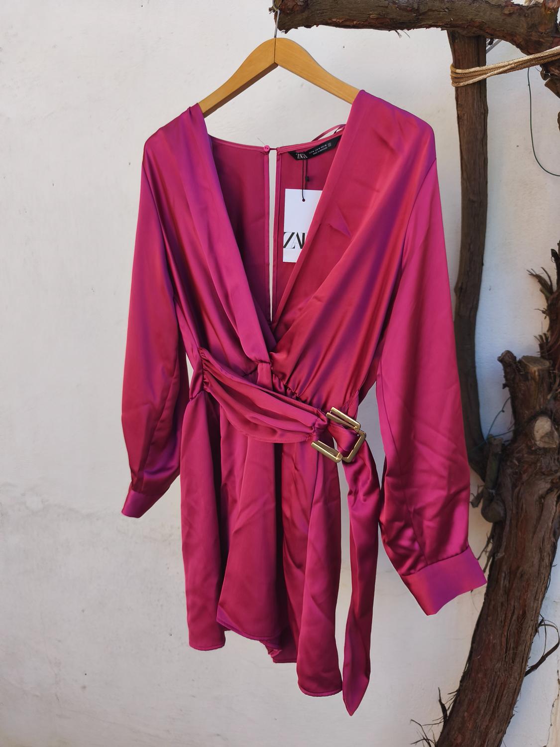 Fuchsia satin dress with belt and gold buckles, hanging on a wooden hanger in front of a white wall. Label visible.