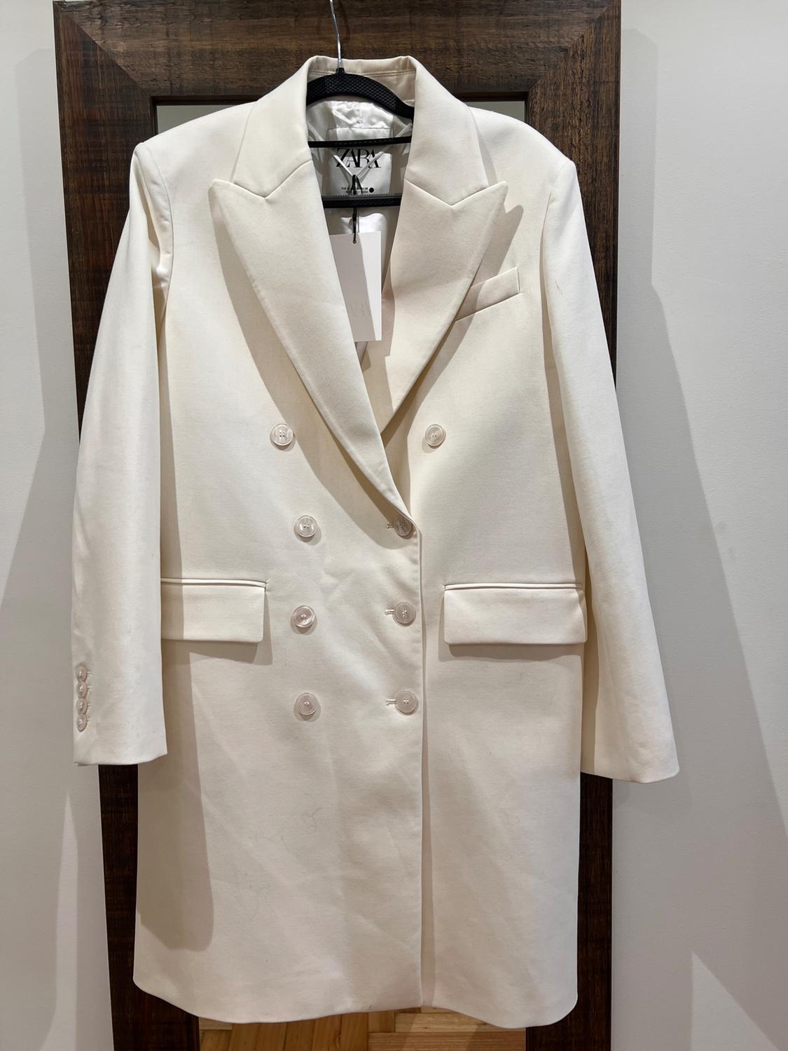 White double-breasted coat hanging on a hanger, with wide lapels and front pockets, against a wooden background.