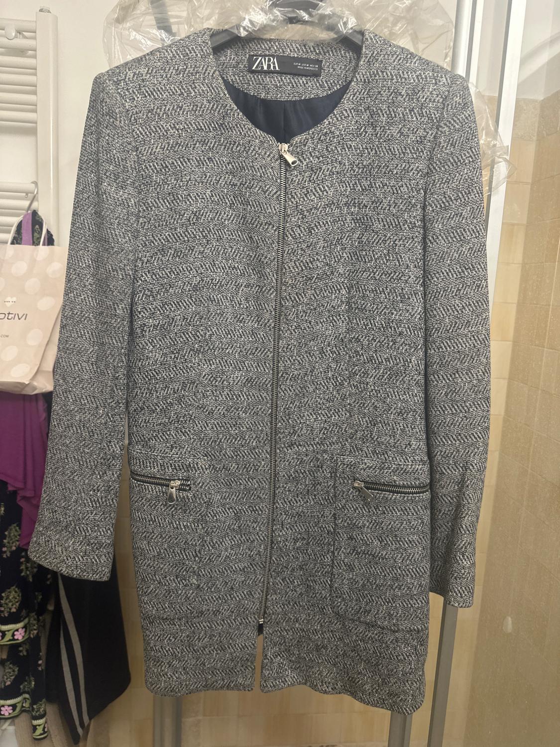 Grey coat with zip and fastening pockets, hanging on a hanger. Visible label: "ZARA". Background with bags and clothes hanging up.