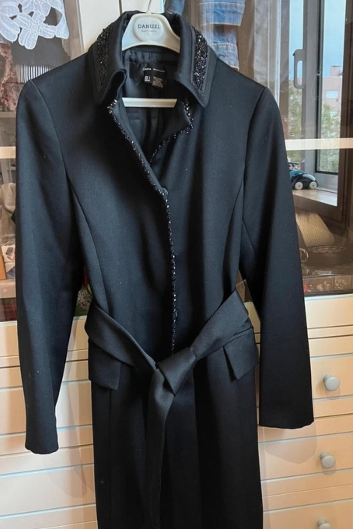 Front view of black coat with adorned collar and lapel, belt, and side pockets.