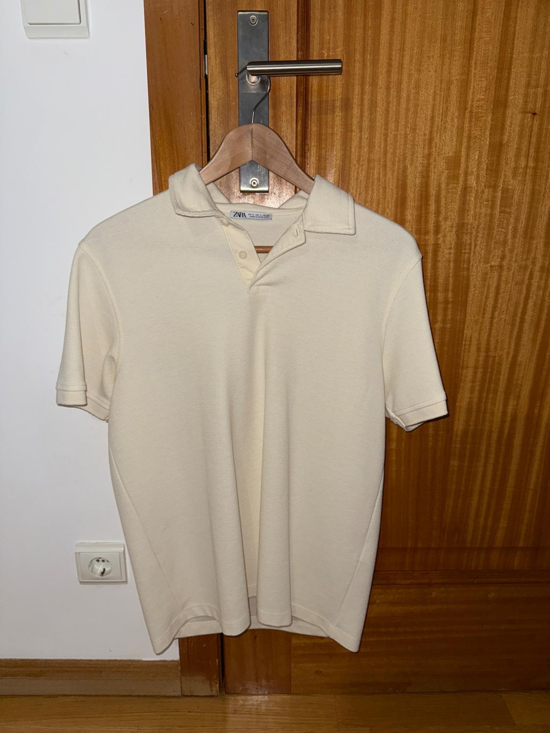 Beige polo shirt hanging on a wooden hanger over a wooden door. Label visible on the collar. Plug socket on the white wall.
