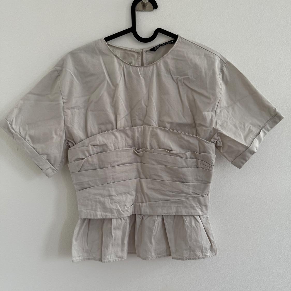 Beige short-sleeve blouse with pleated design on the torso and ruffle detail on the hem, hanging on a black hanger against a white wall.