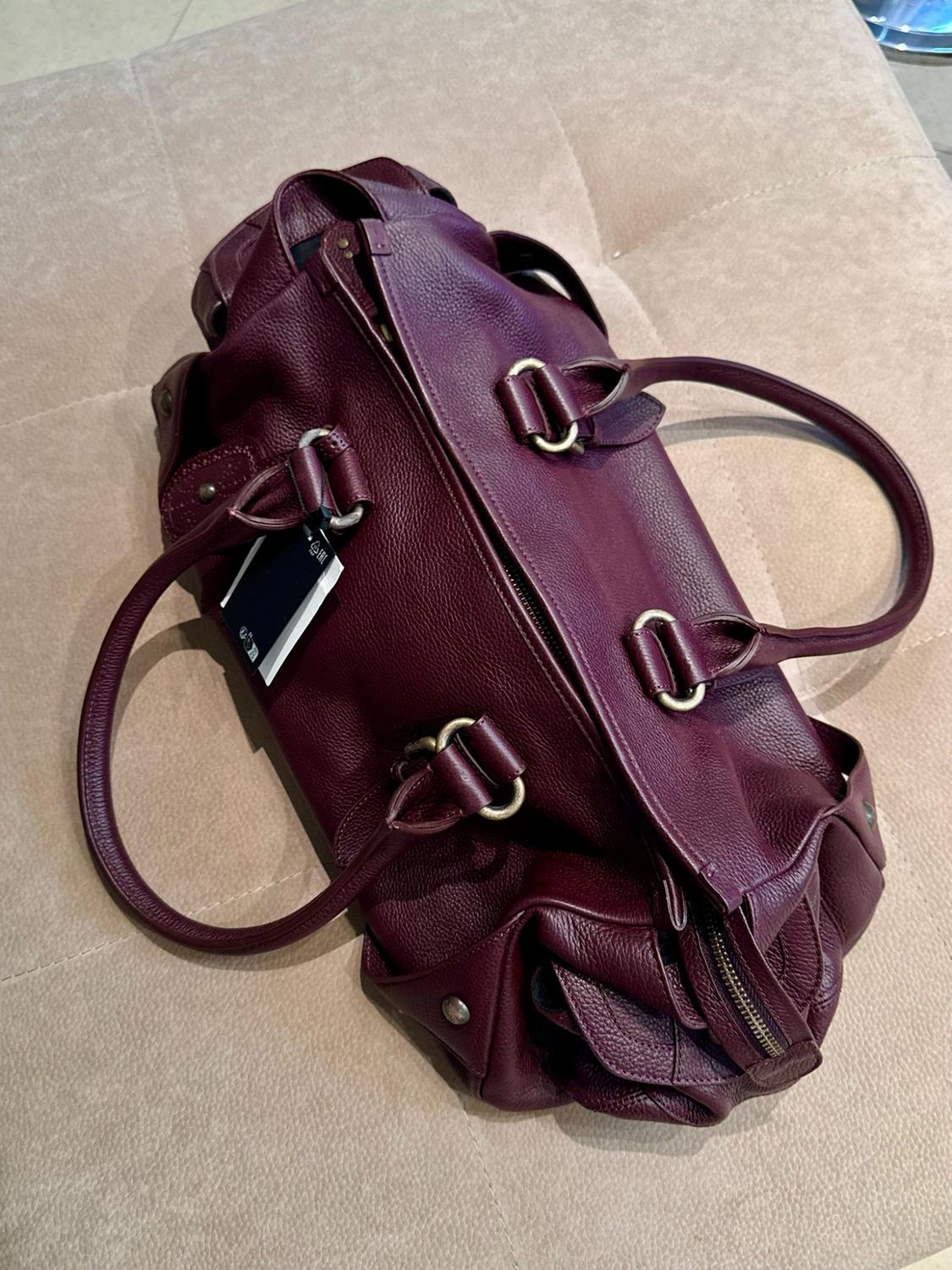 Large burgundy textured leather bag with top handles, metal details and a tag, on a beige surface.