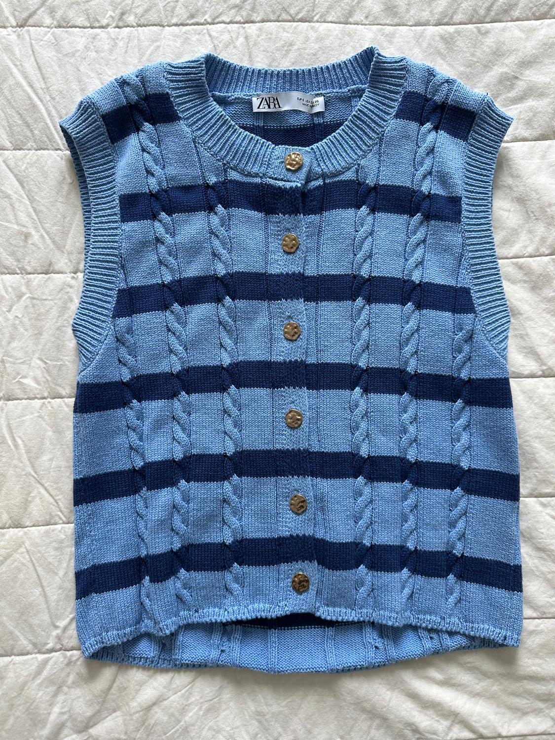 Cable-knit waistcoat with light and dark blue stripes and gold buttons. Label with the text ZARA. Light fabric background.