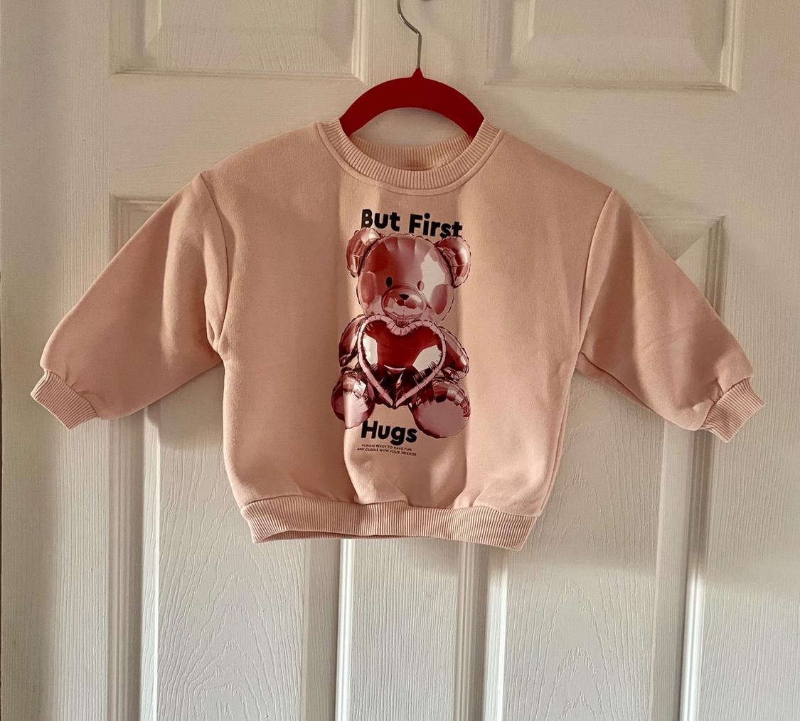 Light pink sweatshirt hanging on a door, with a balloon bear print and "But First Hugs" text on the front.