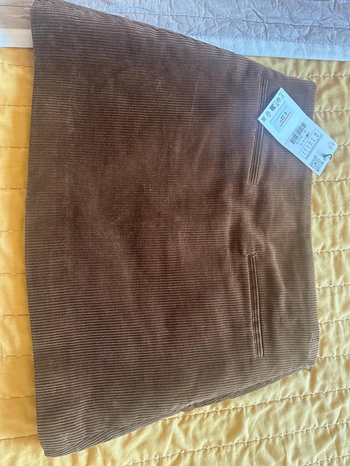 Short brown corduroy skirt with pockets and a label bearing the text S €22.95, laid out on a yellow bedspread.