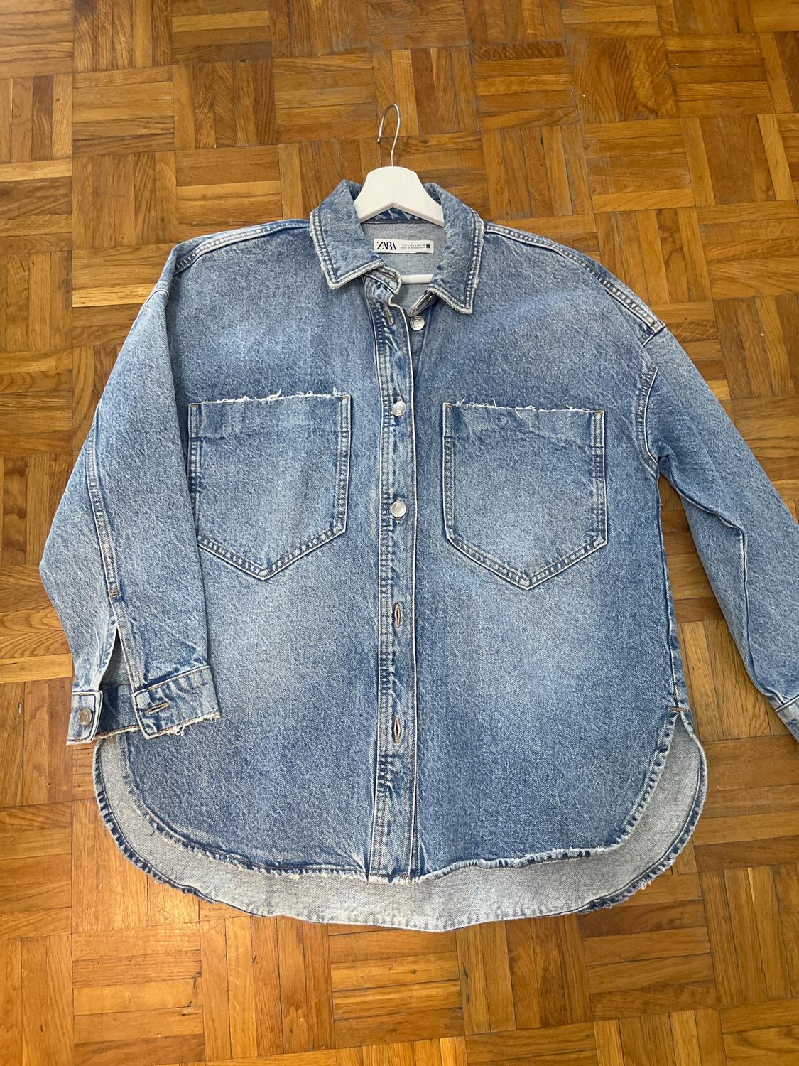 Light blue denim shirt with two front pockets, hanging on a hanger over a wooden floor. Visible tag: Zara.