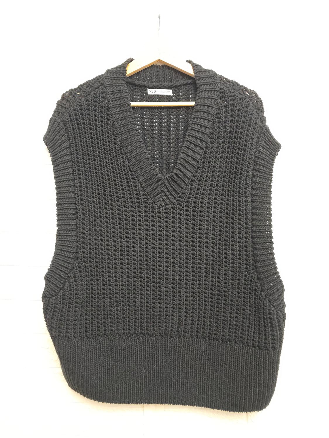 Black knitted V-neck vest hanging on a wooden hanger against a white background. Label visible on the neck.