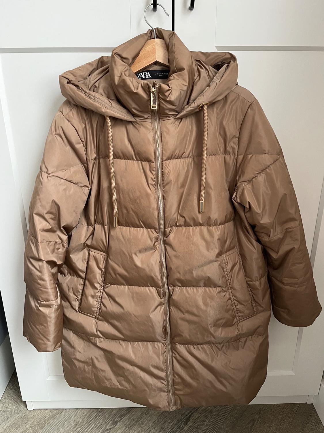 Brown padded coat with a hood and zip fastening, hanging on a hanger in front of a white wardrobe.