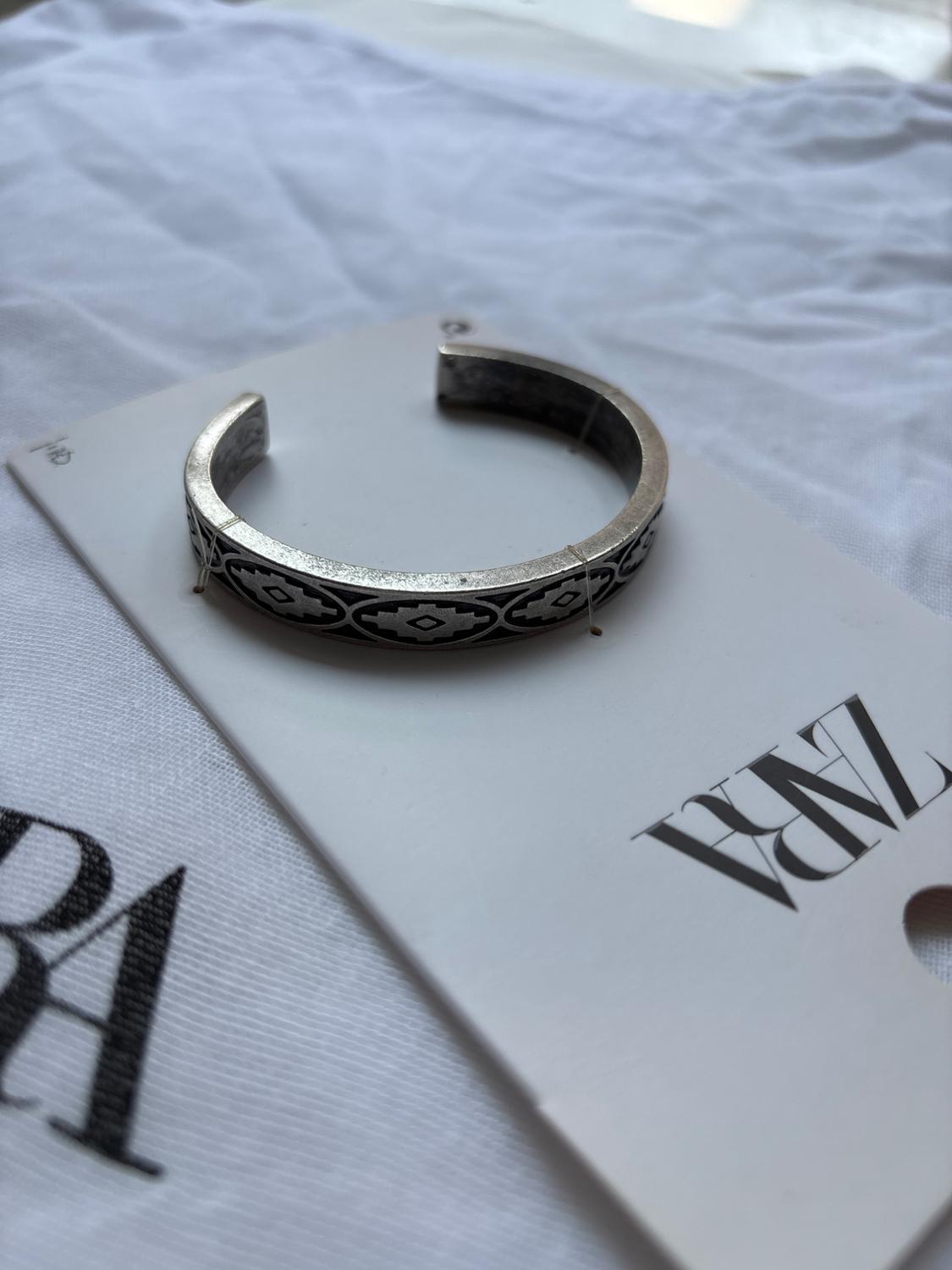 Silver bracelet with geometric design on white card with "ZARA" text on wrinkled white fabric background.