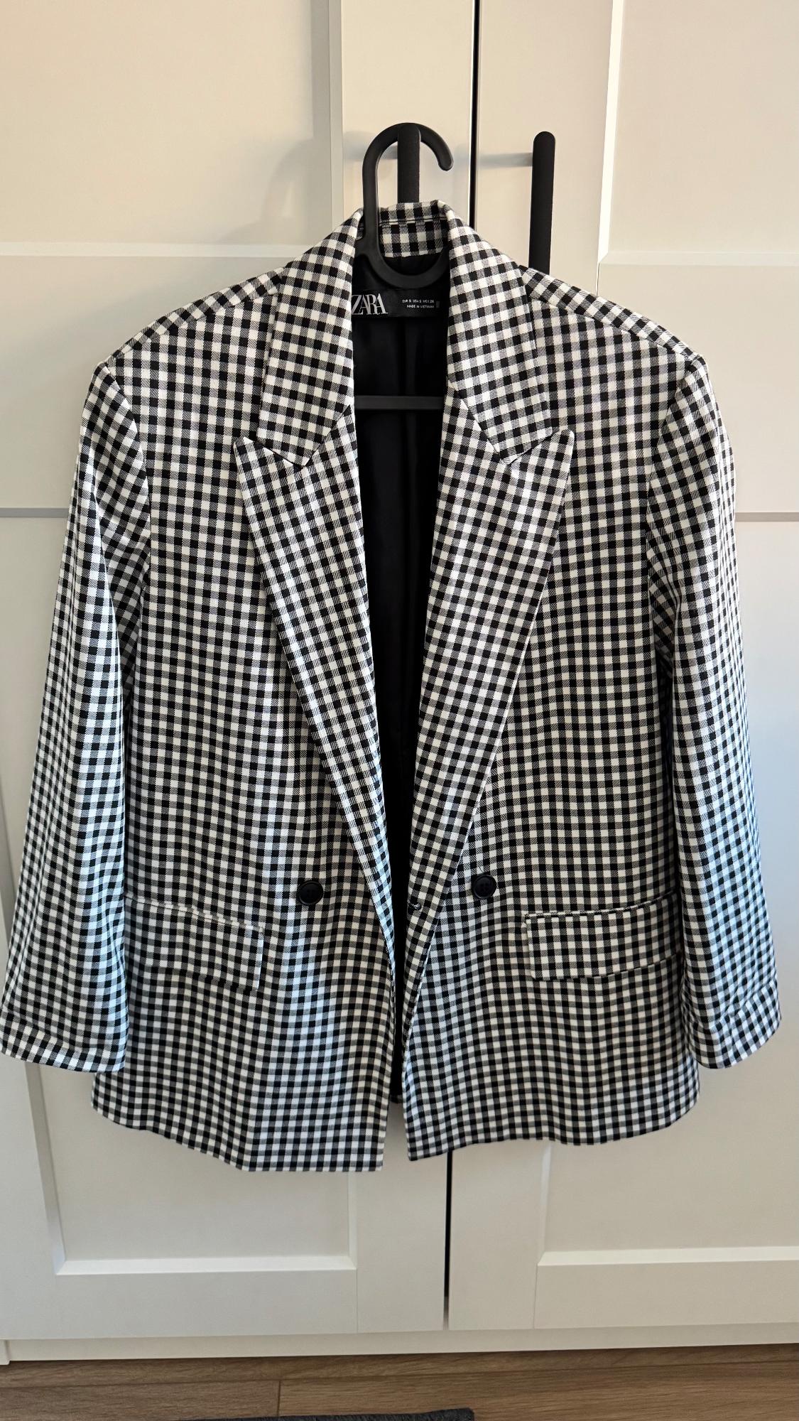 Black and white checked jacket hanging on a black coat hanger in front of a white door. Label visible: Zara.