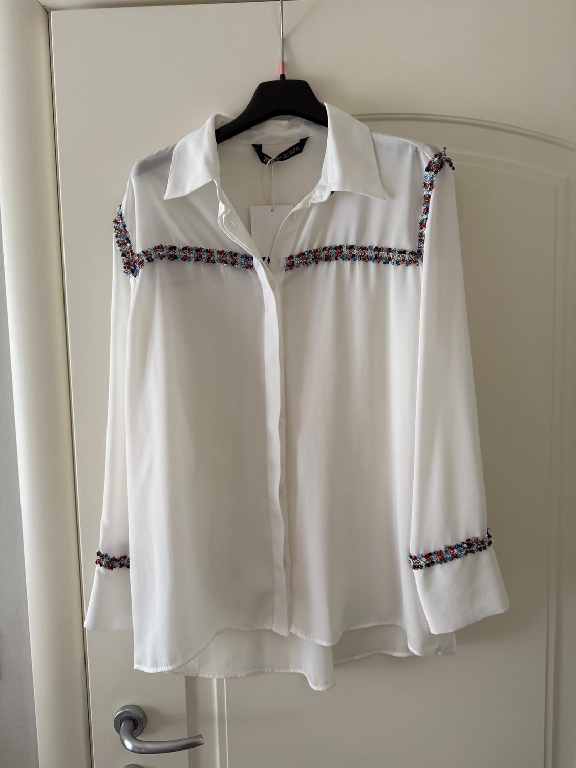 White blouse hanging on a black hanger, with colourful embroidered details on the shoulders and sleeves, over a white door.