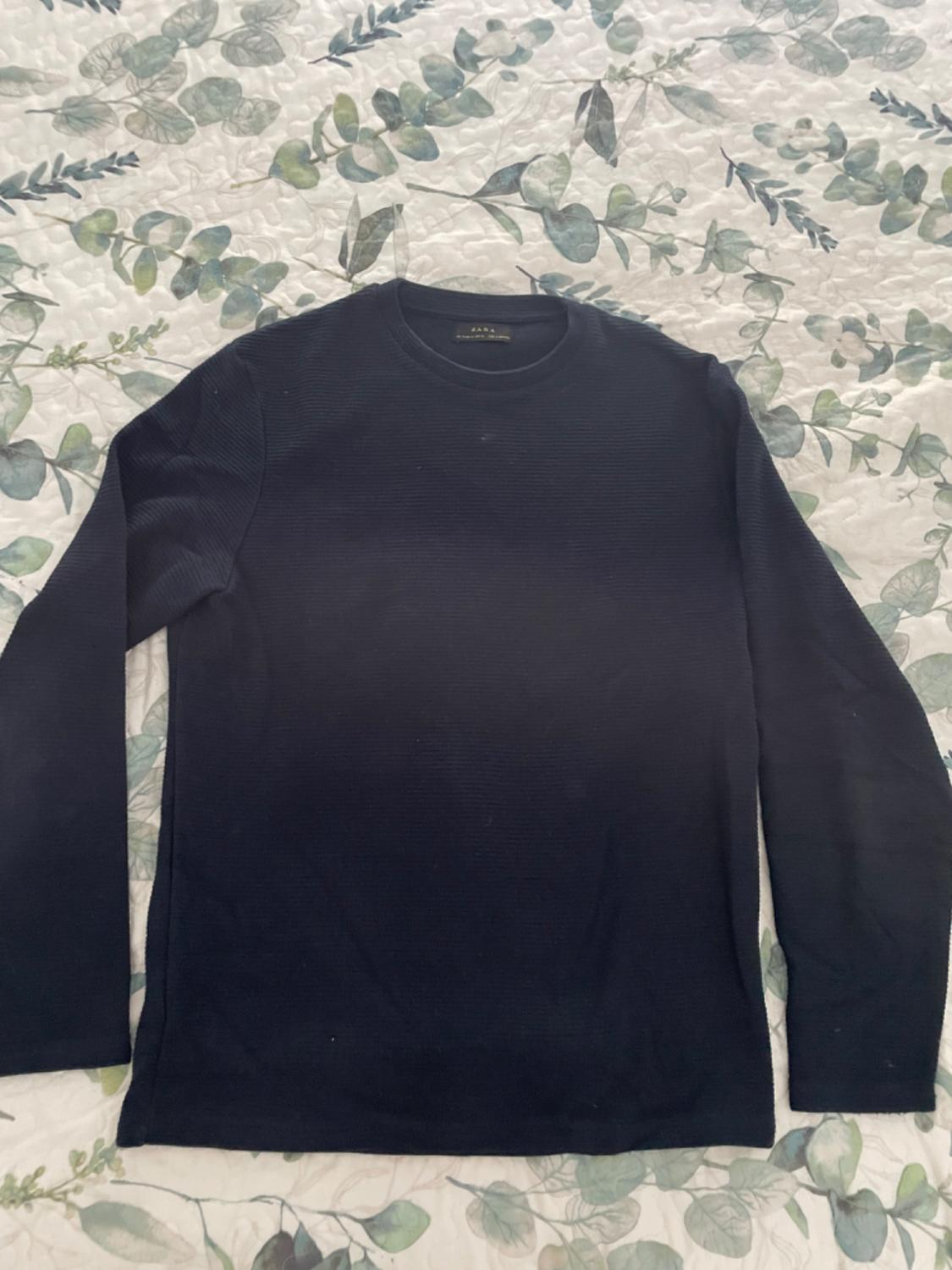 Black long-sleeved jumper on a bedspread with a green leaf print. Label visible on the collar.