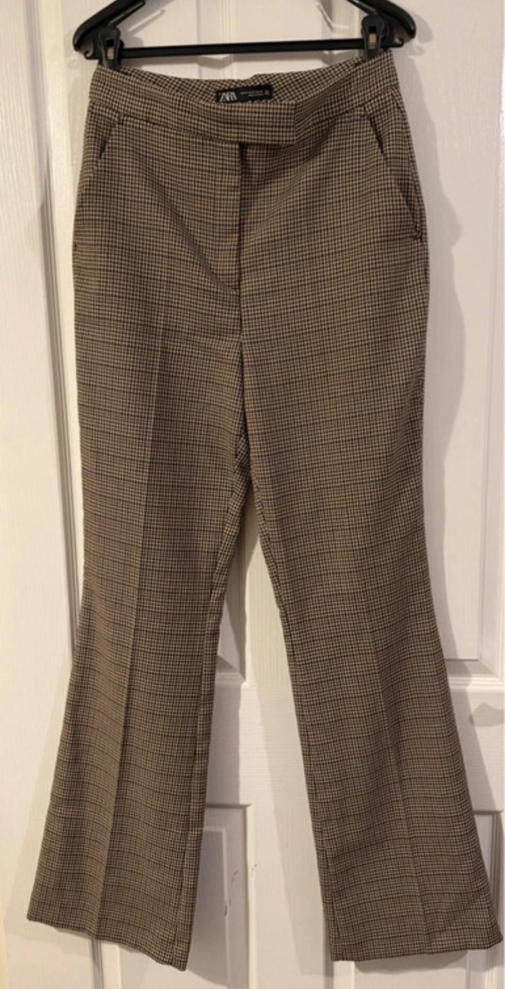Brown checked trousers hanging on a black hanger over a white door. Label visible on the waistband.