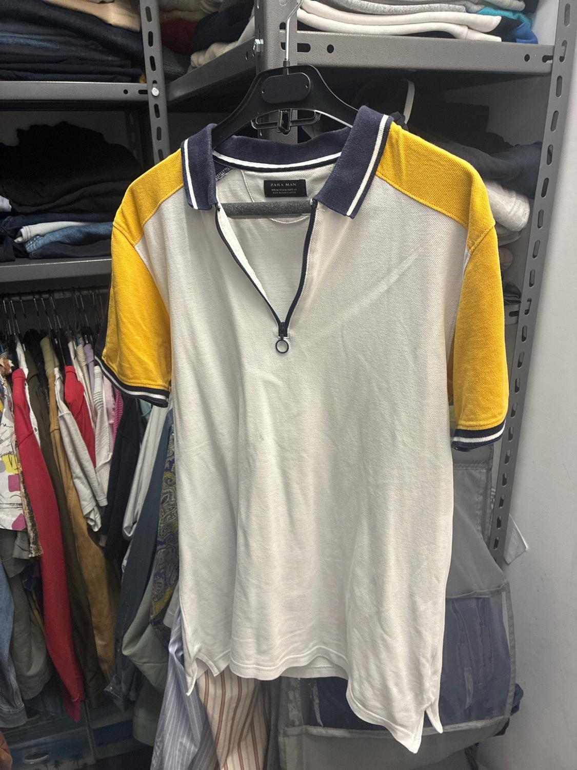 Λευκό polo με κίτρινα μανίκια και μπλε γιακά με φερμουάρ, κρεμασμένο σε μια ντουλάπα γεμάτη ρούχα. Στην ετικέτα αναγράφεται ZARA MAN.