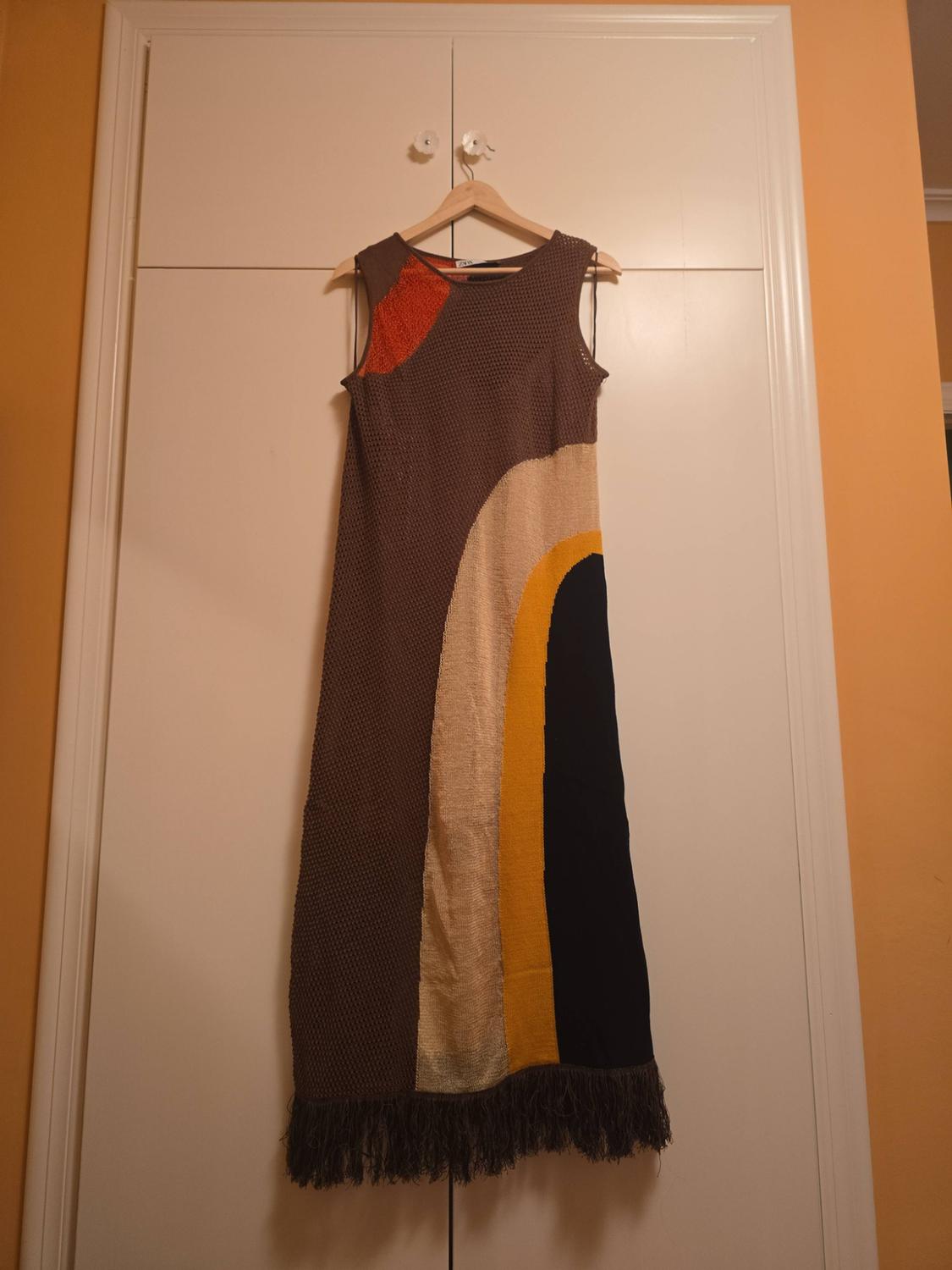 Long sleeveless knitted dress hanging in a wardrobe. Colour-block design in brown, orange, beige, yellow and black, with a fringed hem.