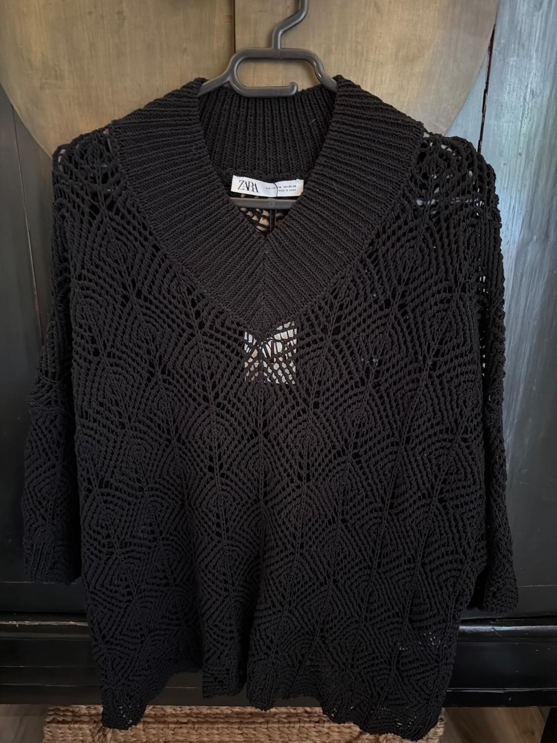 Black open-knit jumper with geometric design hanging on a hanger. Visible label with text "ZARA". Dark wooden background.