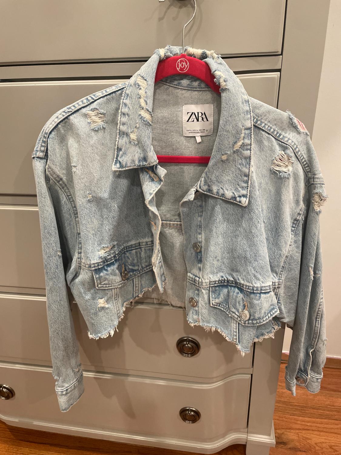 Distressed denim jacket from ZARA hanging on a red hanger in front of a gray cabinet.