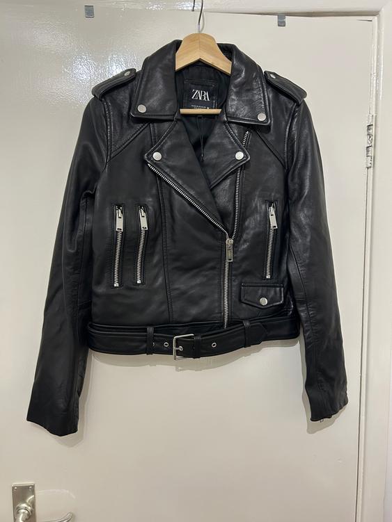 Black leather jacket with zips and belt, hanging on a wooden hanger. Visible label: ZARA. White background.