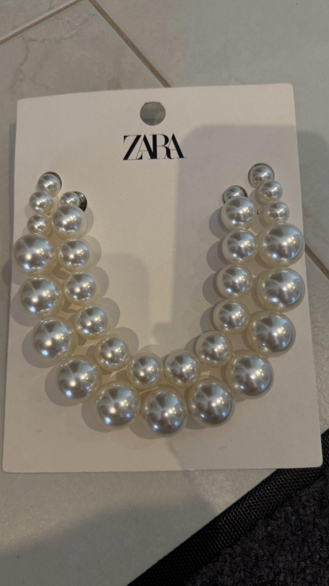 White pearl necklace of varying sizes on a cardboard backing with the text ZARA, resting on a tiled floor.