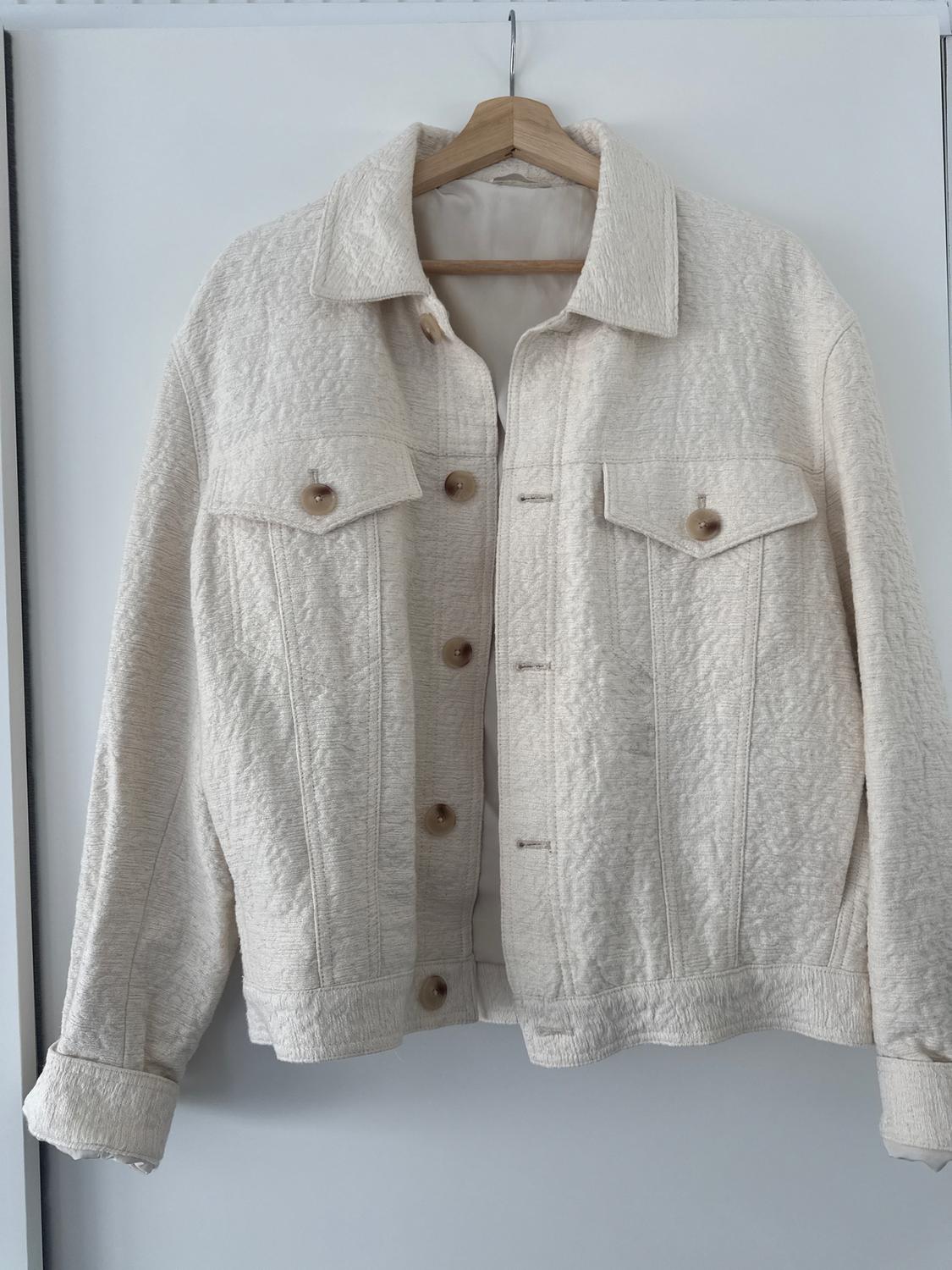 Cream textured fabric jacket hanging on a wooden hanger, with brown buttons and front pockets.