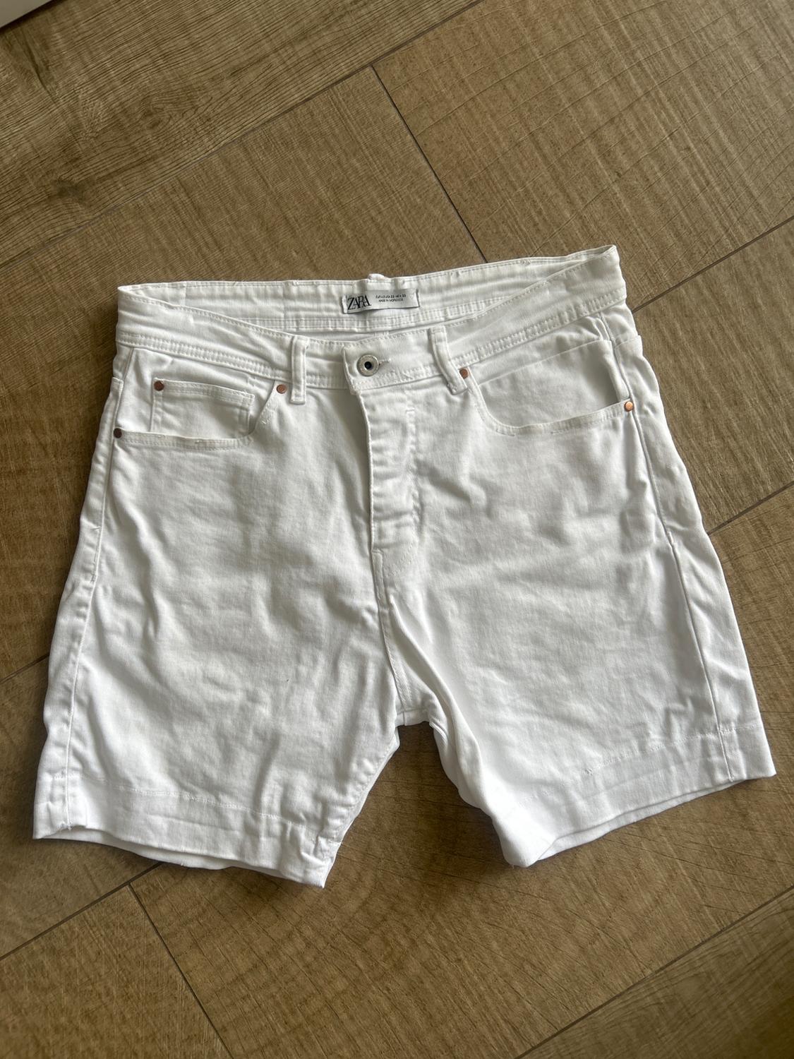 White denim shorts with ZARA label, five pockets and button, laid out on a wooden floor. Casual and relaxed style.