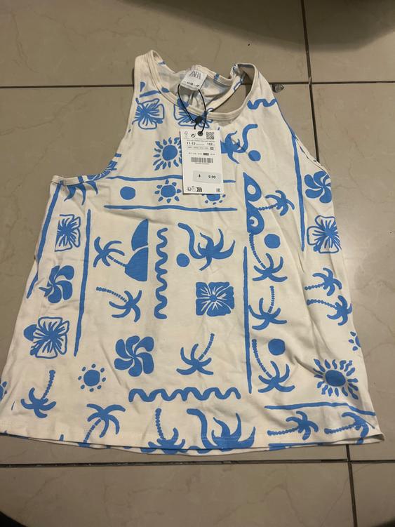 White sleeveless shirt with blue floral and palm tree print. Visible tag with $9.90 price. Beige tile background.