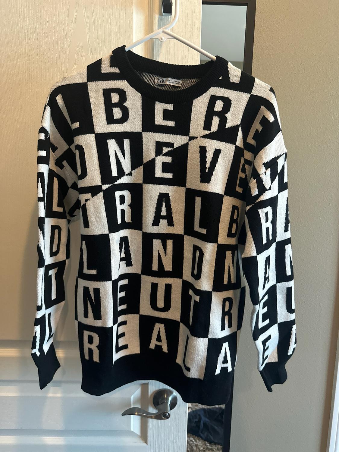 Black-and-white checked jumper with printed capital letters and a ZARA label. It is hanging on a hanger on a white door.