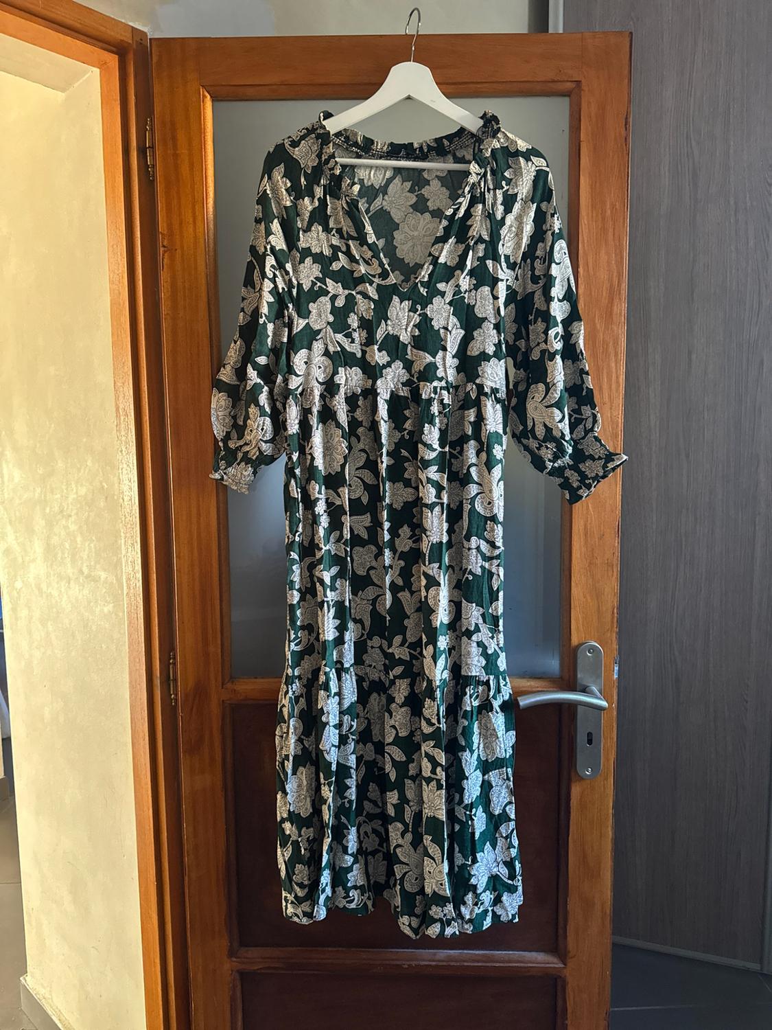 Dark green maxi dress with a white floral print, V-neck and three-quarter sleeves, hanging on a hanger on a wooden door.