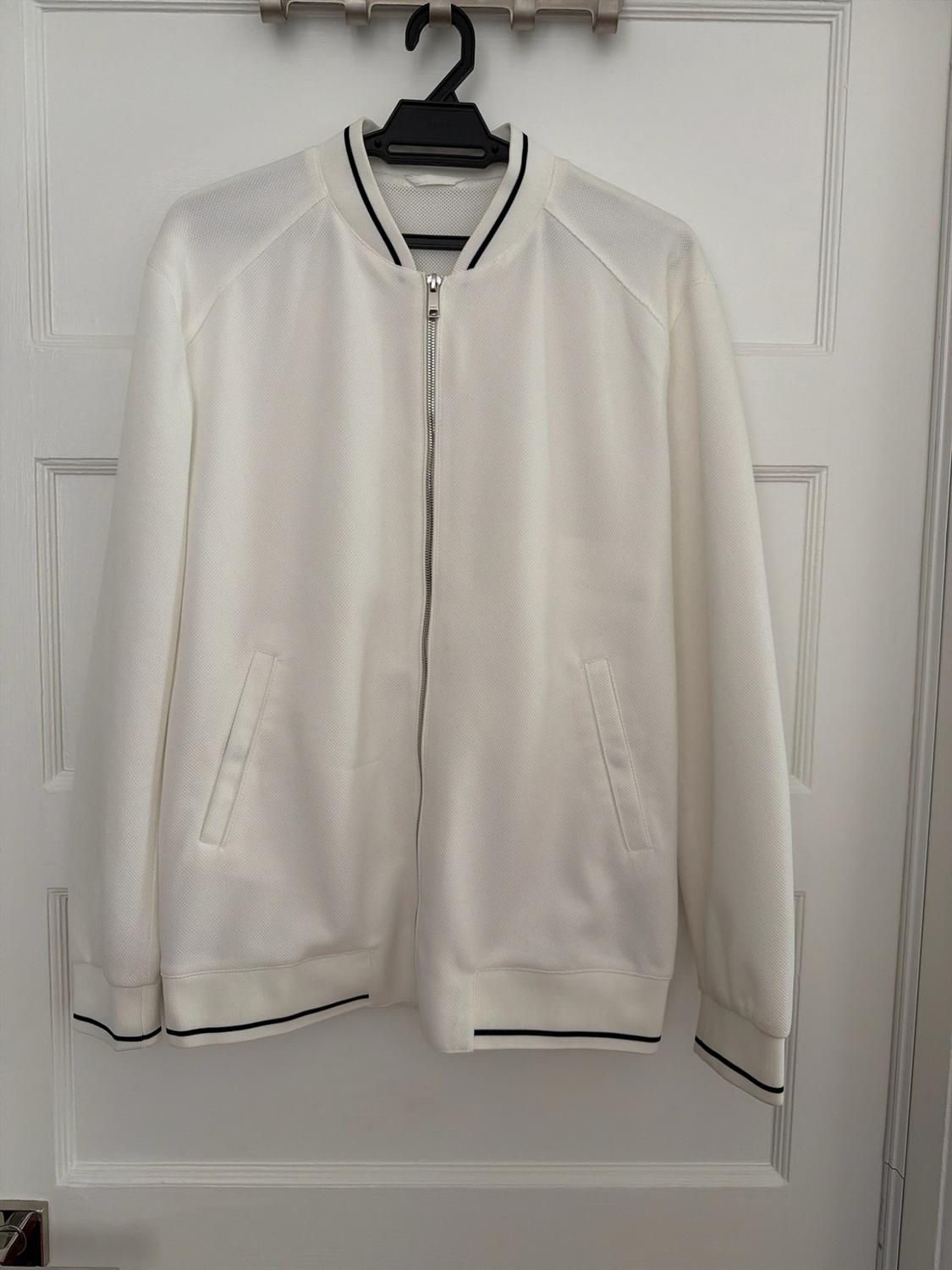 White zippered jacket with black stripes on the collar, cuffs, and hem, hanging on a hanger over a white door.