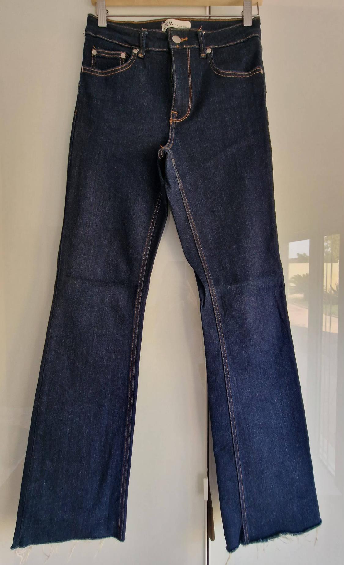 Dark blue denim trousers hanging on a hanger. Contrasting stitching detail, frayed hem and ZARA label at the waist.
