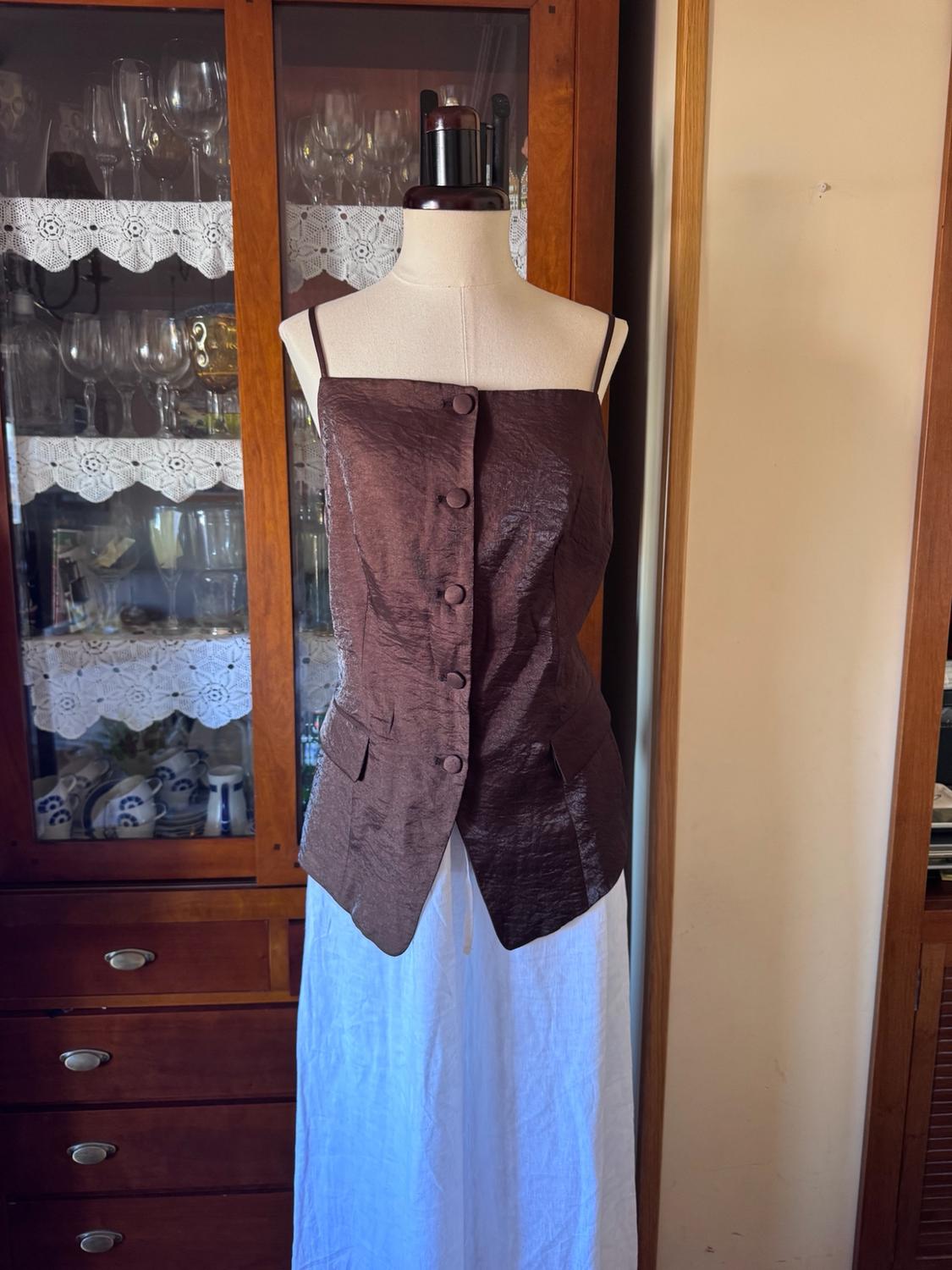 Mannequin wearing a brown textured strappy top with buttons, and a white skirt, next to a wooden display cabinet with glassware.