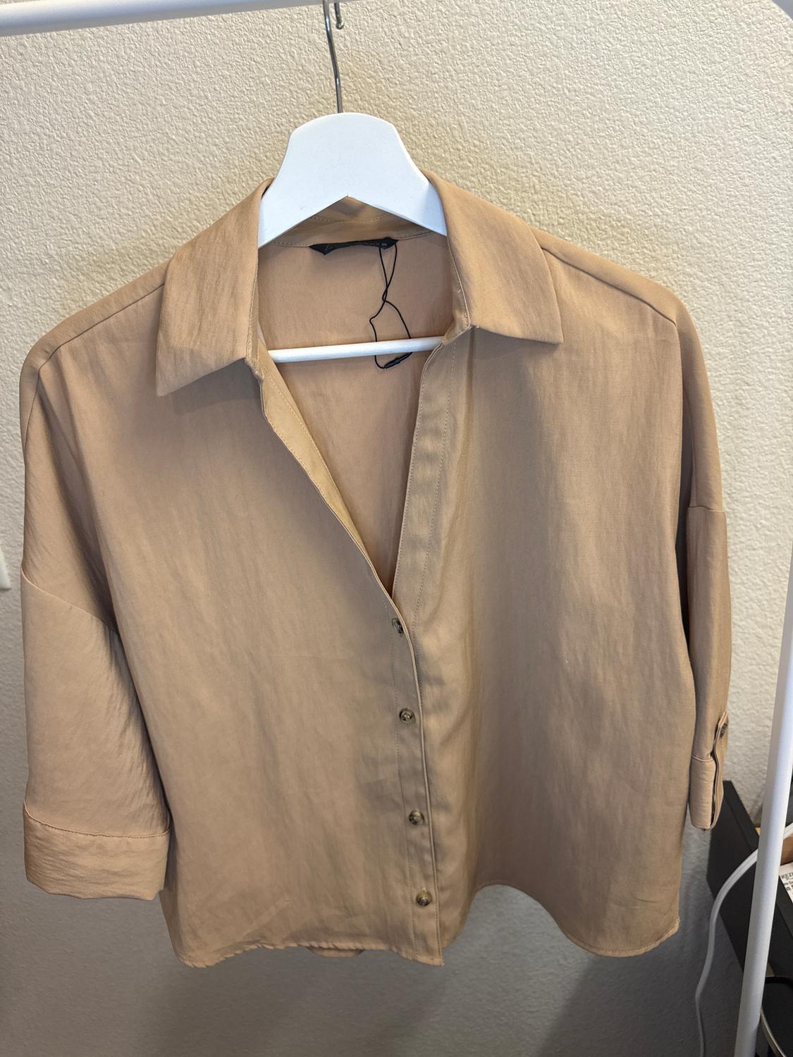 Beige long-sleeved shirt hanging on a white hanger, with front buttons and a classic collar, on a light background.