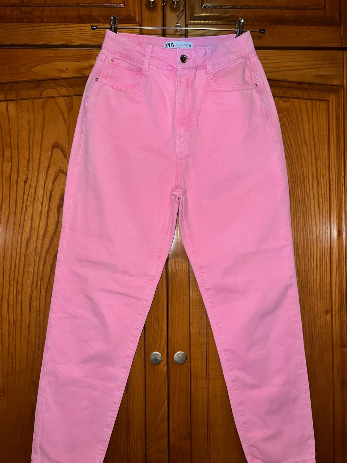 Pink jeans hanging on a metal hanger over wooden wardrobe doors. The inside label reads ZARA.