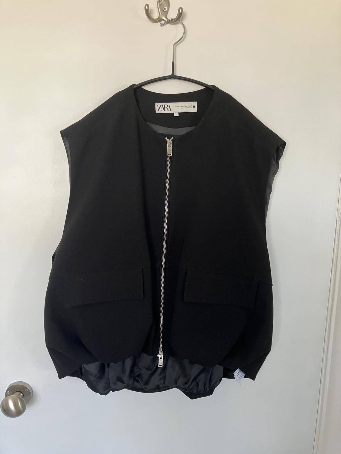 Black sleeveless gilet with a front zip and large pockets, hanging on a hanger over a white door. Visible label: Zara.