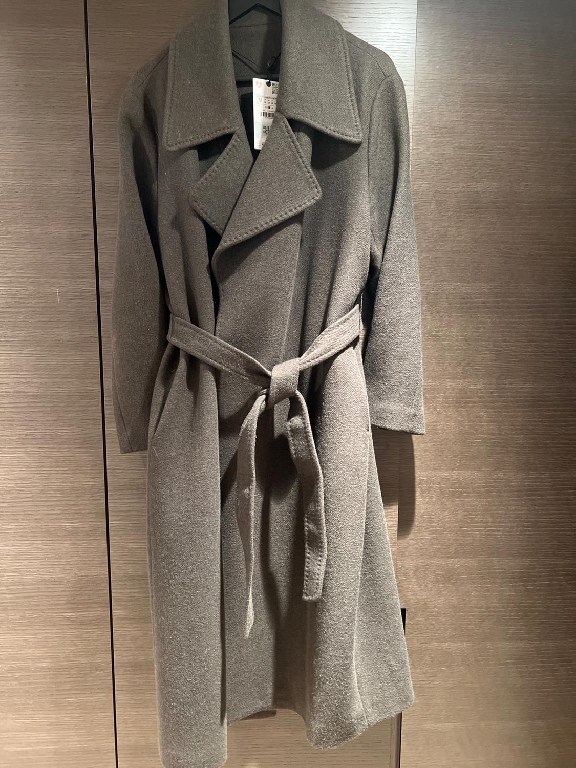 Grey belted coat hanging on a hanger against a wooden background. Visible label on the collar.