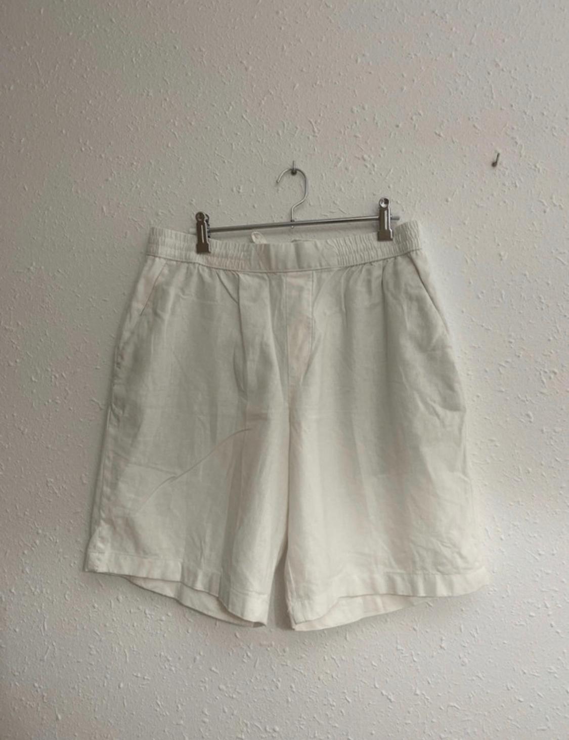 White shorts with an elasticated waistband hanging from a metal hanger on a textured white wall. Relaxed and minimalist style.