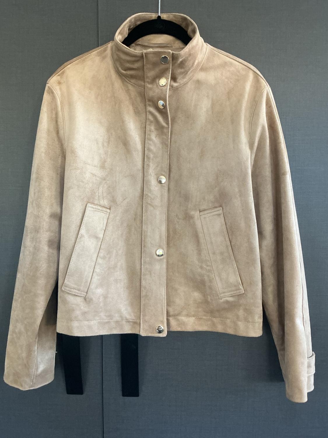 Beige jacket with a high collar, snap buttons, and side pockets, hanging on a black hanger against a dark gray background.