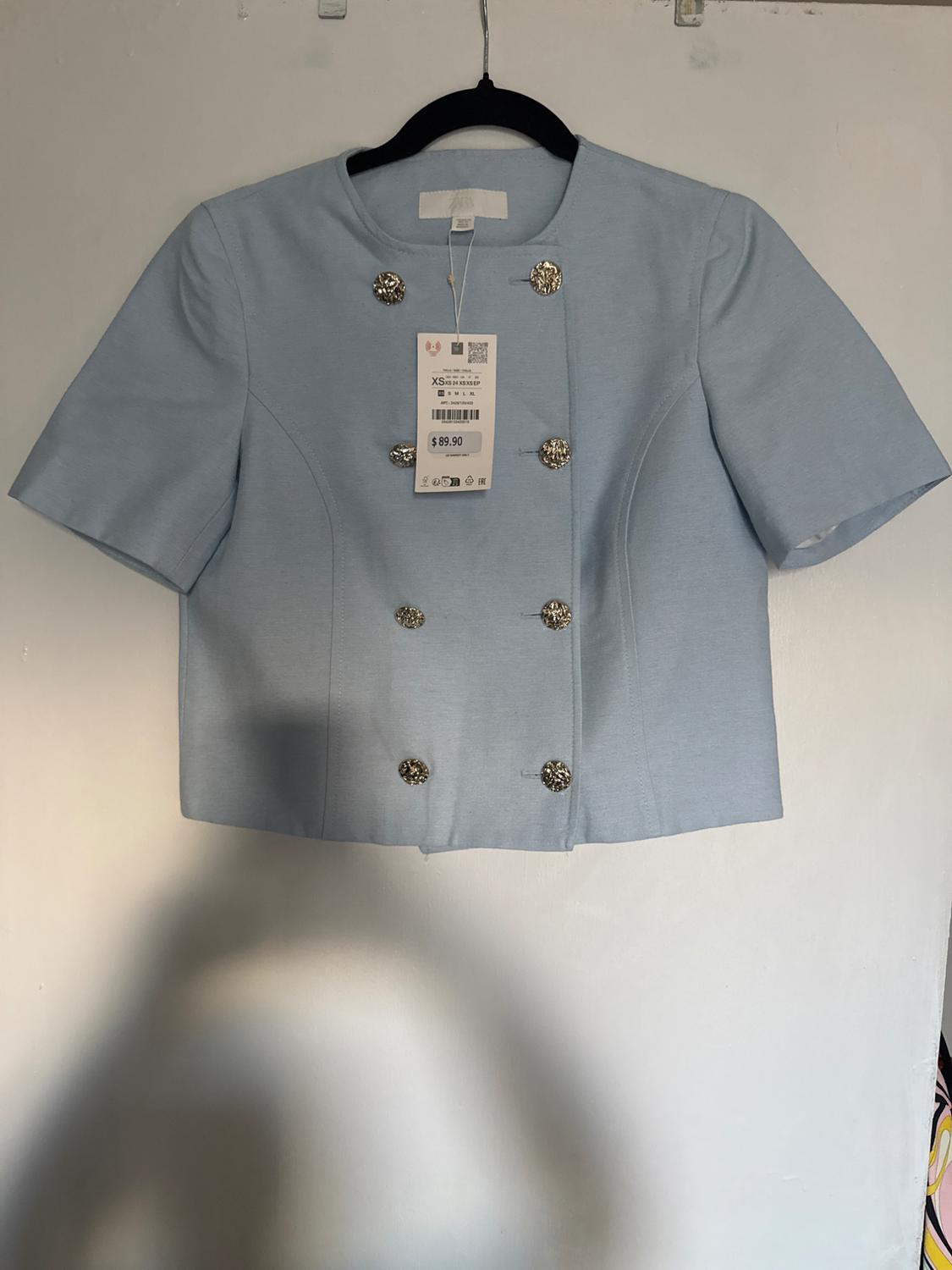 Light blue jacket with decorative buttons, hanging on a black hanger. Visible tag with price of $89.90. White background.