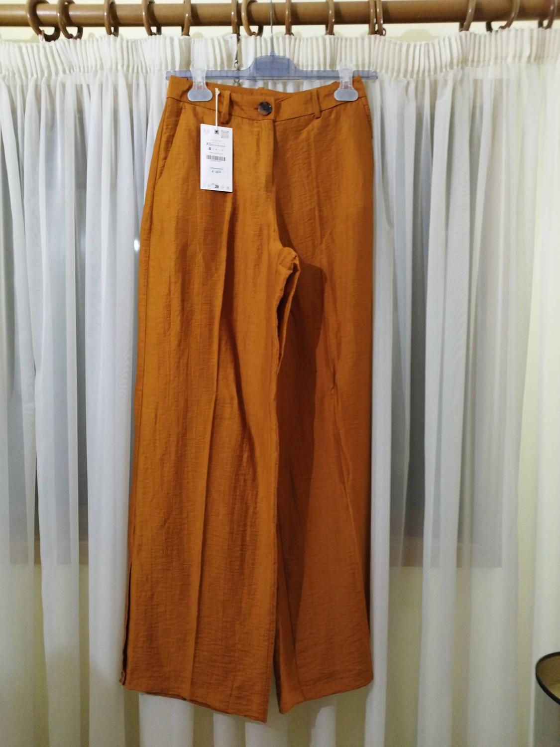 Wide terracotta-coloured trousers with a label, hanging on a hanger in front of white curtains. Their textured fabric and dark button stand out.