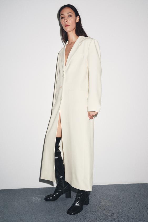 Kombinace dlouhého kabátu v surové barvě s klopami, dlouhými rukávy a paspulkovými kapsami, zkombinovaná s černými botami na podpatku.