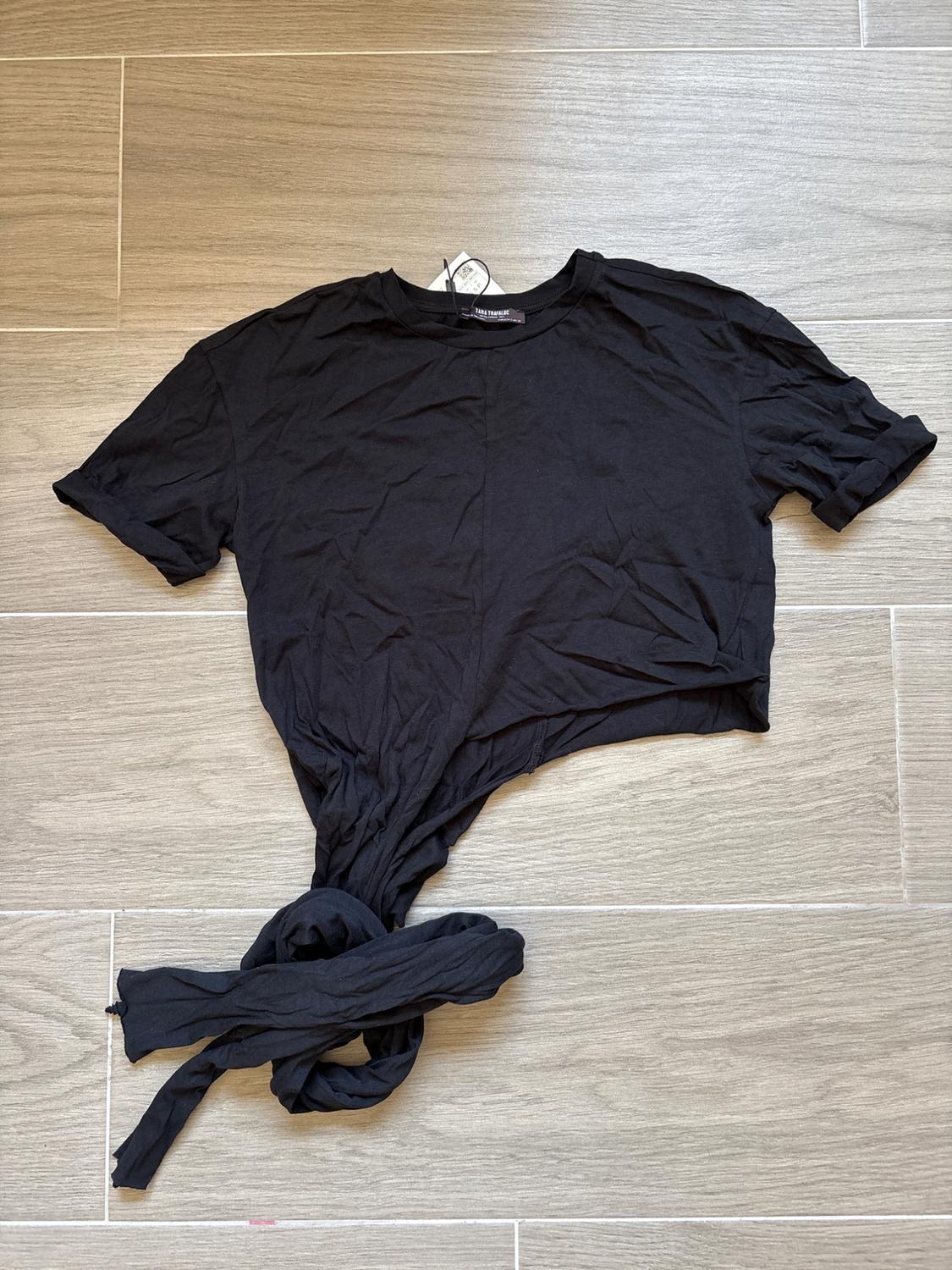 Black short-sleeve T-shirt with a knotted design at the bottom, placed on a light wood-coloured tiled floor.