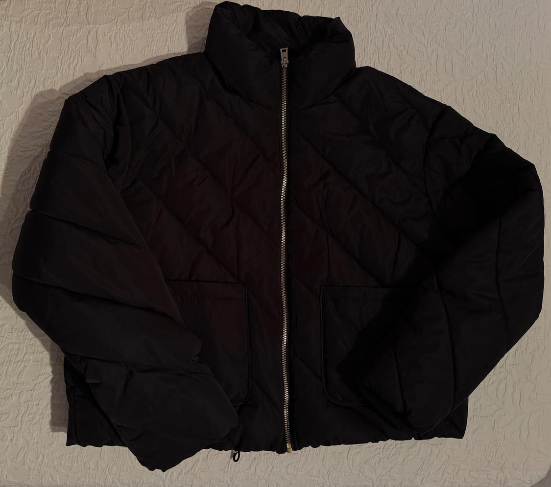 Black padded jacket with front zip on a textured white background. High-neck design and front pockets.
