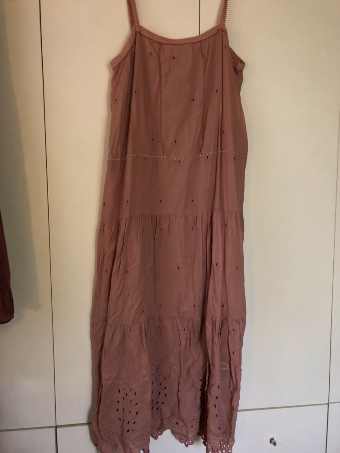 Long, spaghetti-strap dress in a dusky pink colour, with embroidered and perforated detailing on the lower section, hanging on a pale wall.
